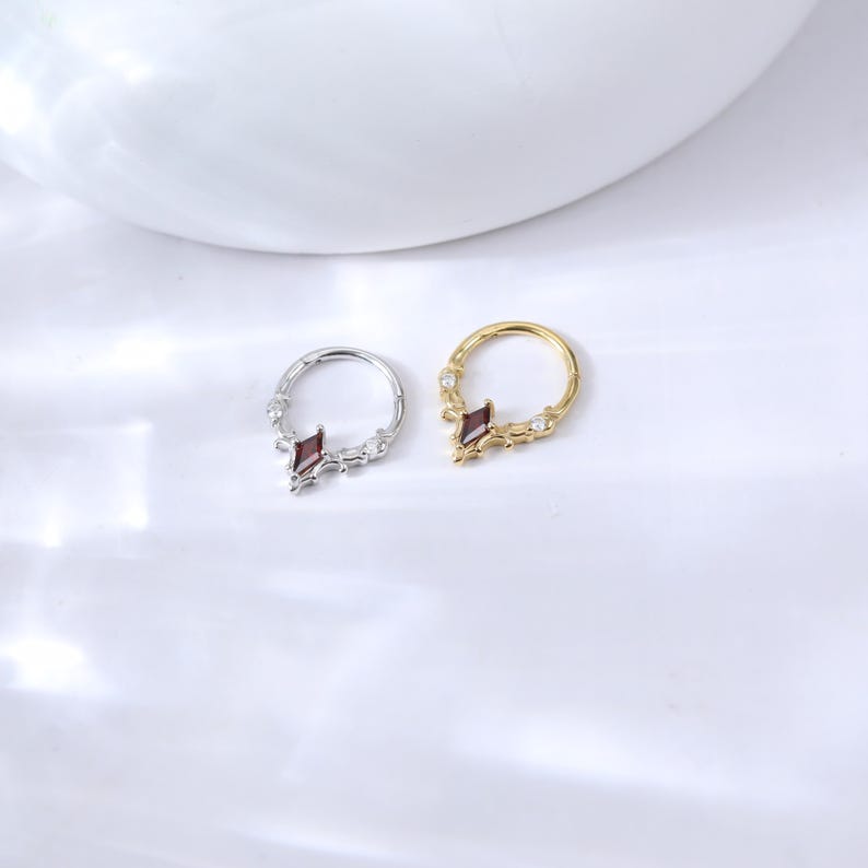 May include: Two nose rings, one silver and one gold, are displayed. Each ring features a central, dark red, diamond-shaped gem and smaller clear gems. The rings are set against a white background.