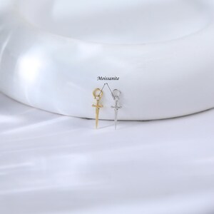 May include: Two small pendant earrings in the shape of daggers. One is gold-colored, the other is silver-colored. Both have a small gemstone at the hilt and are labeled "Moissanite".