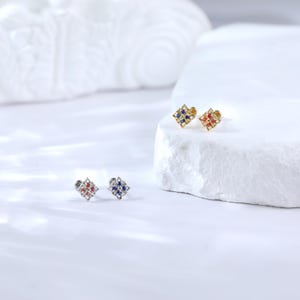 May include: Three pairs of small, square-shaped earrings with a decorative design. The earrings are available in gold, silver, and rose gold with blue and red gemstones.
