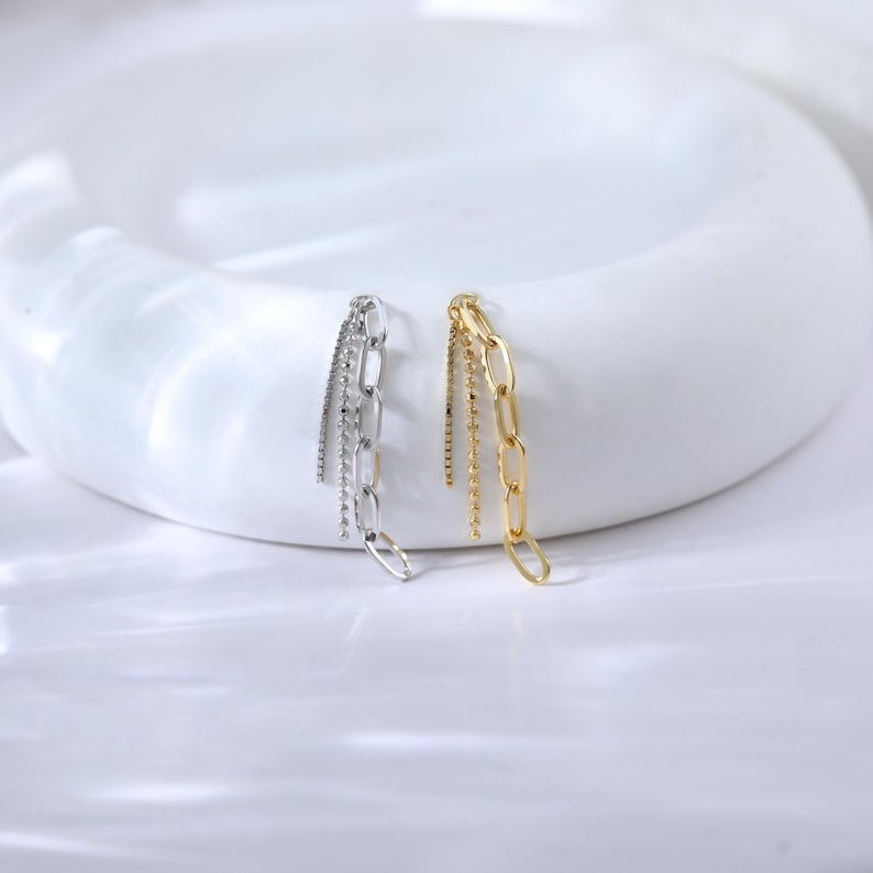 May include: Two pairs of earrings, one silver and one gold, are displayed. The silver pair features a chain design with dangling strands of small beads. The gold pair has a chain design with dangling strands of small beads. Both pairs are on a white surface.