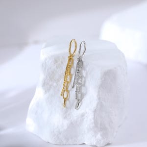 May include: A pair of dangling earrings, one gold-toned and one silver-toned, displayed on a white, textured surface. The gold earring features a chain design with a beaded accent, while the silver earring has a chain and a line of small, clear stones.