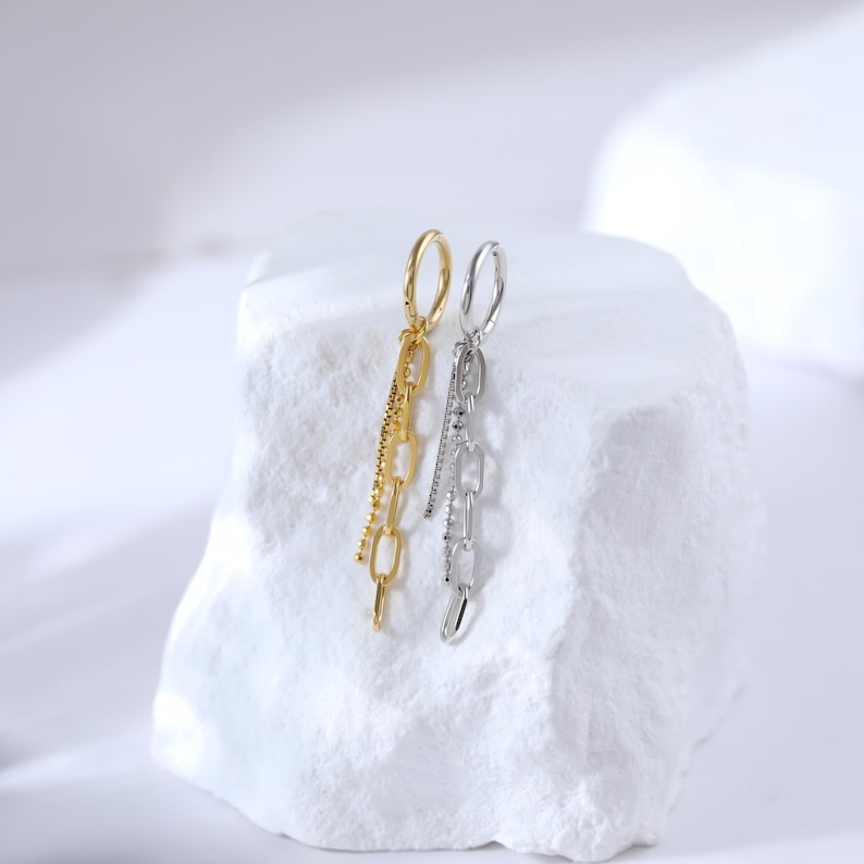 May include: A pair of dangling earrings, one gold-toned and one silver-toned, displayed on a white, textured surface. The gold earring features a chain design with a beaded accent, while the silver earring has a chain and a line of small, clear stones.