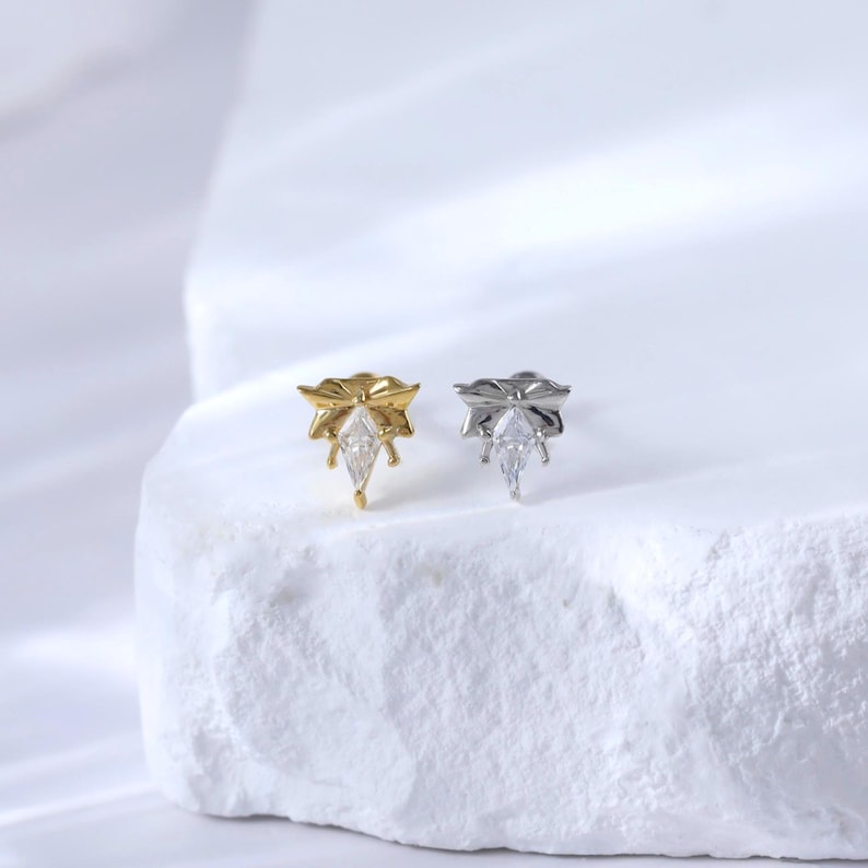 Puede incluir: Dos pendientes de bot&oacute;n, uno dorado y otro plateado, cada uno con una parte superior en forma de lazo y una gema transparente en forma de l&aacute;grima. Los pendientes se muestran sobre una superficie blanca texturizada.