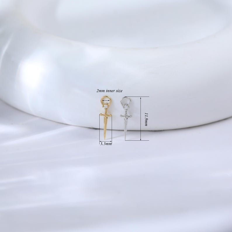 May include: Two small sword-shaped pendants, one gold-colored and one silver-colored, are displayed. The gold pendant has a small clear gem. The silver pendant has a clear gem. The image includes measurements: 2mm inner size, 11.9mm length, and 3.3mm width.