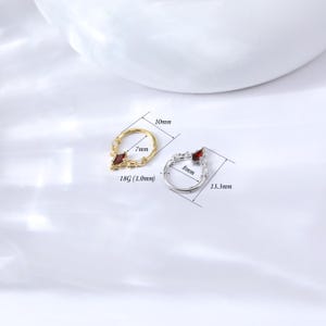 May include: Two nose rings, one gold and one silver, each with a red gemstone. The gold ring measures 10mm by 7mm, and the silver ring measures 13.3mm by 8mm. Both rings have a decorative design.