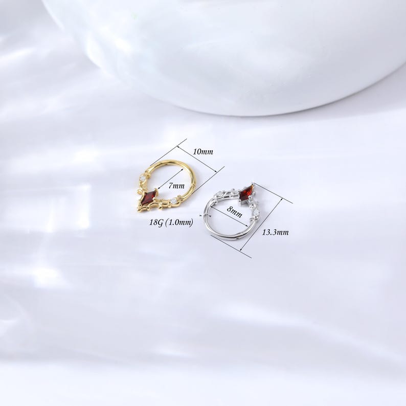 May include: Two nose rings, one gold and one silver, each with a red gemstone. The gold ring measures 10mm by 7mm, and the silver ring measures 13.3mm by 8mm. Both rings have a decorative design.