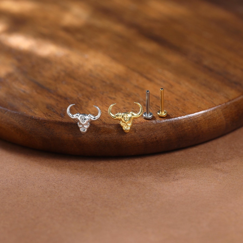 May include: A silver and gold-toned bull head stud earrings and posts are displayed on a wooden surface. The earrings feature detailed bull head designs. The posts are in silver and gold tones. The background is a warm brown.