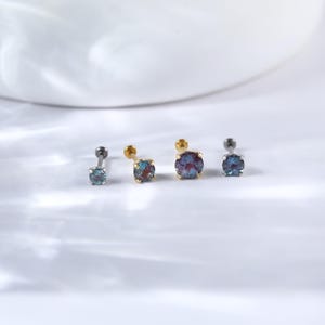 May include: Four silver and gold nose studs with iridescent blue gemstones. The studs are arranged in a row on a white surface.