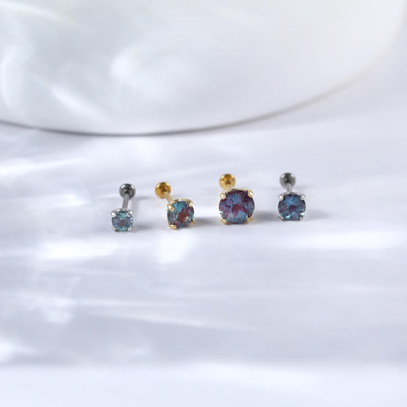May include: Four silver and gold nose studs with iridescent blue gemstones. The studs are arranged in a row on a white surface.