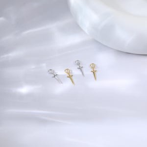 May include: Four small dagger pendants, two silver and two gold, are arranged on a white surface. Each pendant features a detailed hilt and blade design. The pendants are likely for jewelry making or as charms.