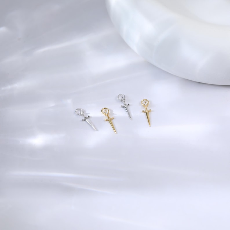 May include: Four small dagger pendants, two silver and two gold, are arranged on a white surface. Each pendant features a detailed hilt and blade design. The pendants are likely for jewelry making or as charms.