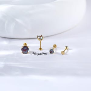 May include: Three gold nose rings with a round, multi-coloured gemstone. The gemstone is a light blue, green, and purple colour. The text "Alexandrite" is written below the rings.