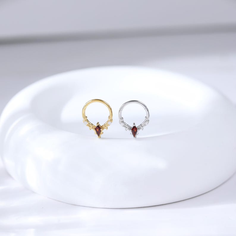 May include: Two small, decorative rings. One is gold-toned, the other silver-toned. Each ring features a central, teardrop-shaped, red gemstone and smaller white gemstones. The rings are displayed on a white, curved surface.