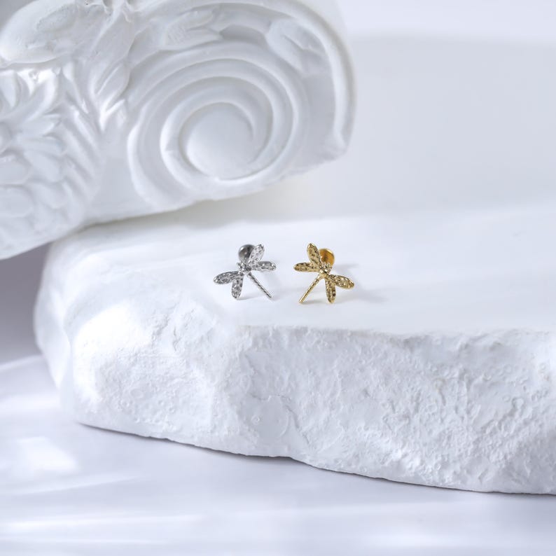 Pu&ograve; includere: Due orecchini a forma di libellula, uno argento e uno oro, su uno sfondo bianco.