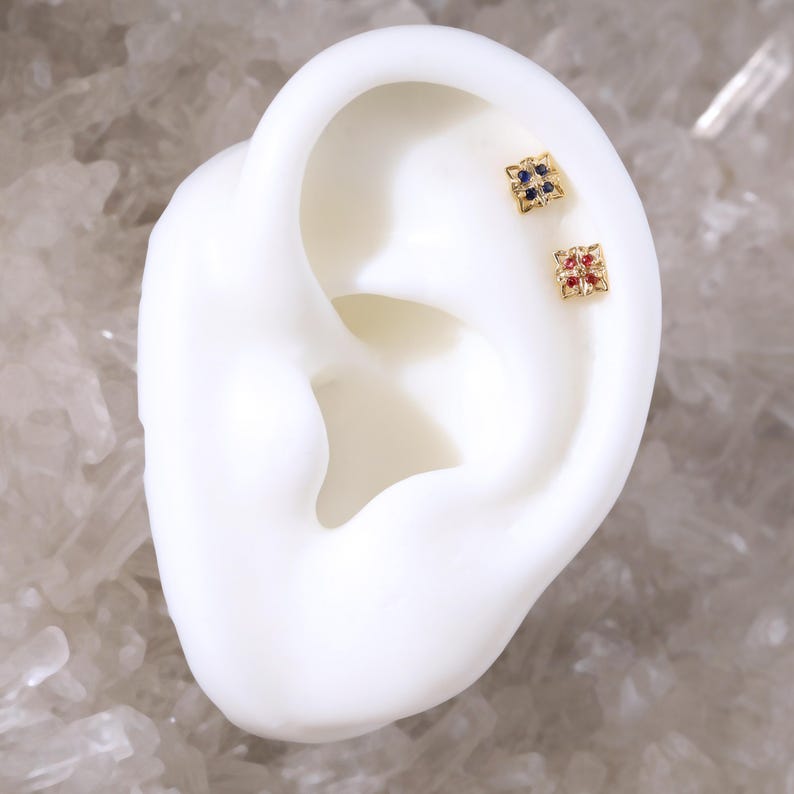 May include: Two small gold earrings with a square design. One earring has blue gemstones and the other has red gemstones.