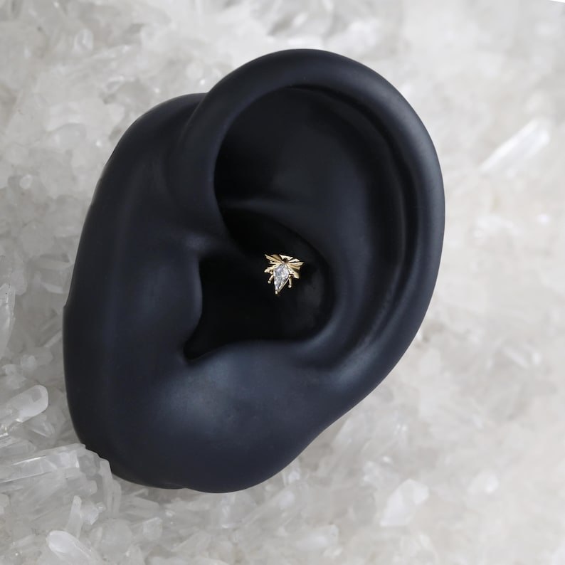 Puede incluir: Primer plano de un piercing de oreja dorado. El piercing presenta un peque&ntilde;o dise&ntilde;o ornamentado con una gema clara central y detalles dorados. El piercing est&aacute; colocado sobre una oreja negra, con un fondo blanco y transparente.