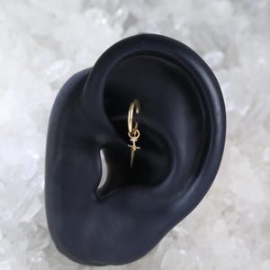 May include: A gold hoop earring with a small dagger pendant. The dagger has a pointed tip and a small gemstone near the top. The earring is shown in a dark ear model against a white background.