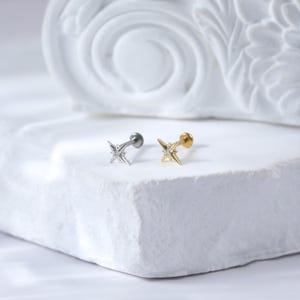 May include: Two silver and gold star-shaped body jewellery pieces with a single clear stone in the centre of each star. The jewellery is on a white surface.