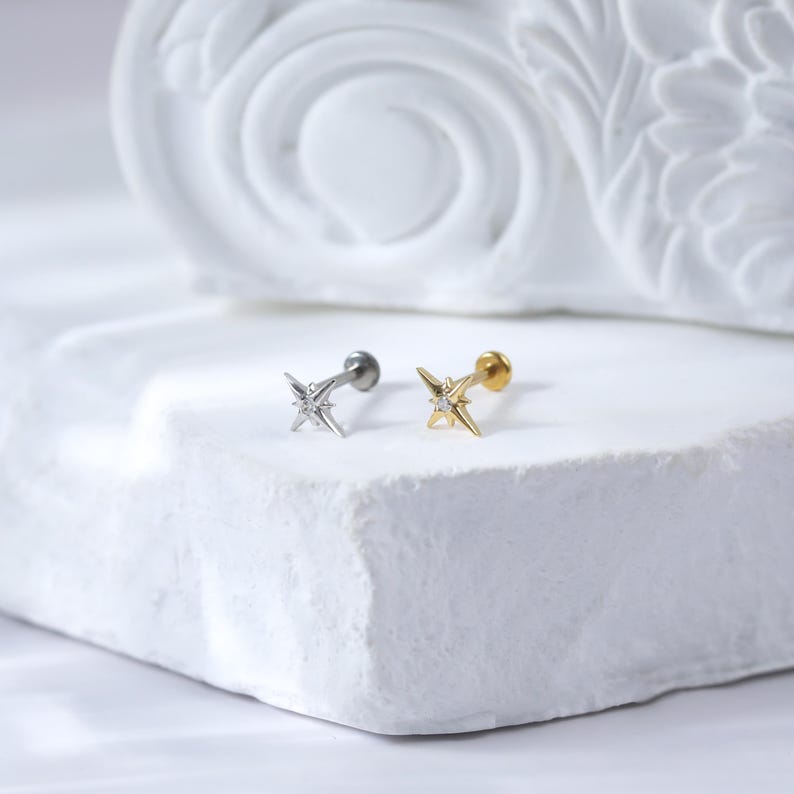 May include: Two silver and gold star-shaped body jewellery pieces with a single clear stone in the centre of each star. The jewellery is on a white surface.