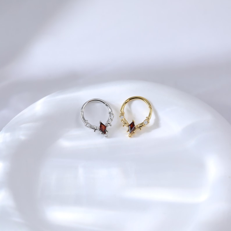 May include: Two hoop earrings, one silver and one gold, each featuring a central, red, diamond-shaped gemstone. The silver earring has a decorative design with clear accents, while the gold earring has clear gemstones.