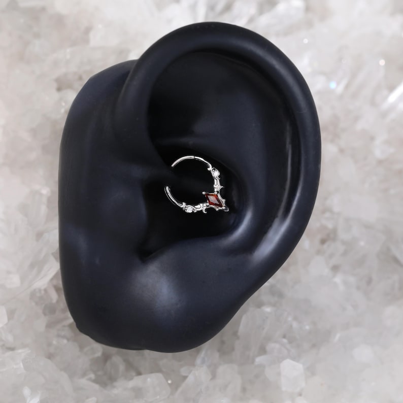 May include: A silver septum ring with a small, red, diamond-shaped gem. The ring is adorned with small, decorative accents. The jewelry is shown in a black ear model against a white, textured background.