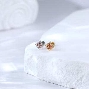 May include: Two small, square-shaped earrings with a delicate, intricate design. The earrings are adorned with red gemstones and are available in gold and silver.
