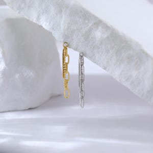 May include: Two dangling earrings, one gold-toned and one silver-toned, hang against a white background. The gold earring features a combination of chain styles, while the silver earring has a delicate chain design. The earrings are a modern jewelry accessory.