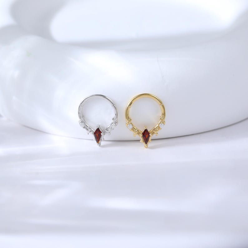May include: Two nose rings, one silver and one gold, each featuring a central red gemstone in a marquise cut. The rings have decorative accents with small, clear stones. The jewelry is displayed on a white surface.