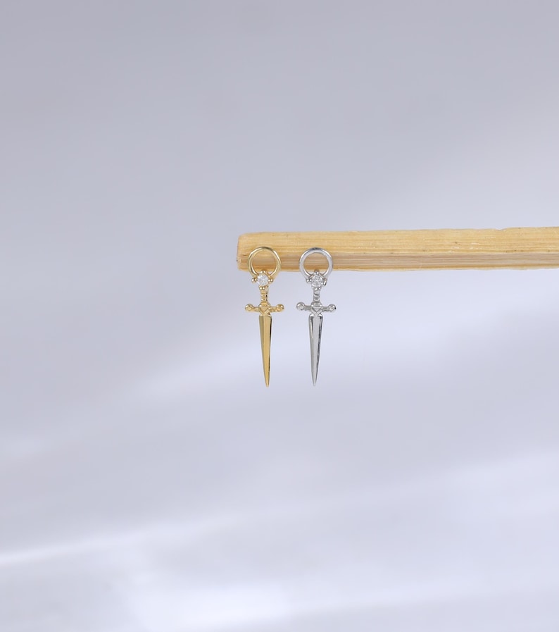 May include: Two small dagger-shaped earrings, one gold-toned and one silver-toned, hang from a wooden dowel. The earrings have a loop at the top and a decorative hilt. The gold earring is on the left, and the silver earring is on the right.