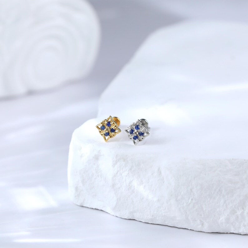 May include: Two small square-shaped earrings with a geometric design and blue gemstones. The earring on the left is gold and the earring on the right is silver.
