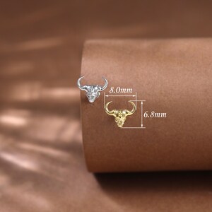 May include: Two bull head-shaped stud earrings, one silver and one gold, are displayed on a brown surface. The silver earring measures 8.0mm, and the gold earring is 6.8mm in length. The earrings have a detailed skull design with curved horns.