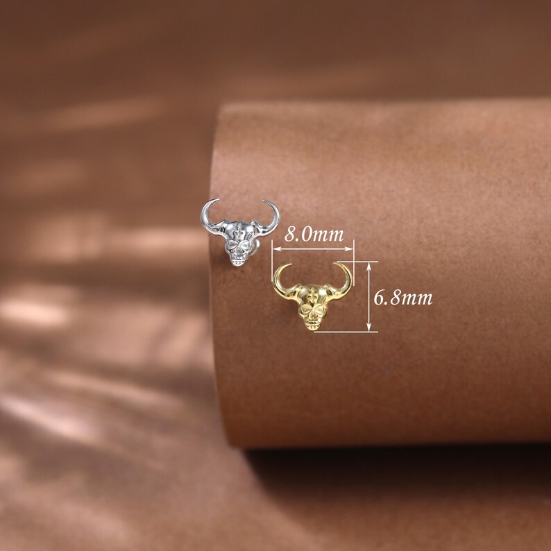 May include: Two bull head-shaped stud earrings, one silver and one gold, are displayed on a brown surface. The silver earring measures 8.0mm, and the gold earring is 6.8mm in length. The earrings have a detailed skull design with curved horns.