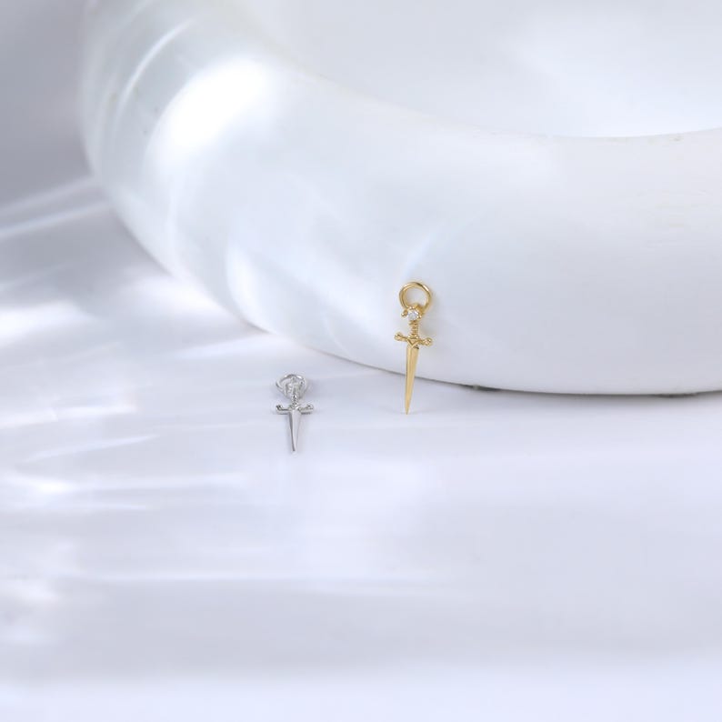 May include: Two small pendant charms. One is silver-colored, shaped like a rose with a cross. The other is gold-colored, shaped like a sword with a loop at the top. Both charms have small, clear accent stones.
