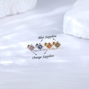 May include: Three different colored square-shaped earrings with a small stud post. The earrings are labeled "Blue Sapphire" and "Orange Sapphire".