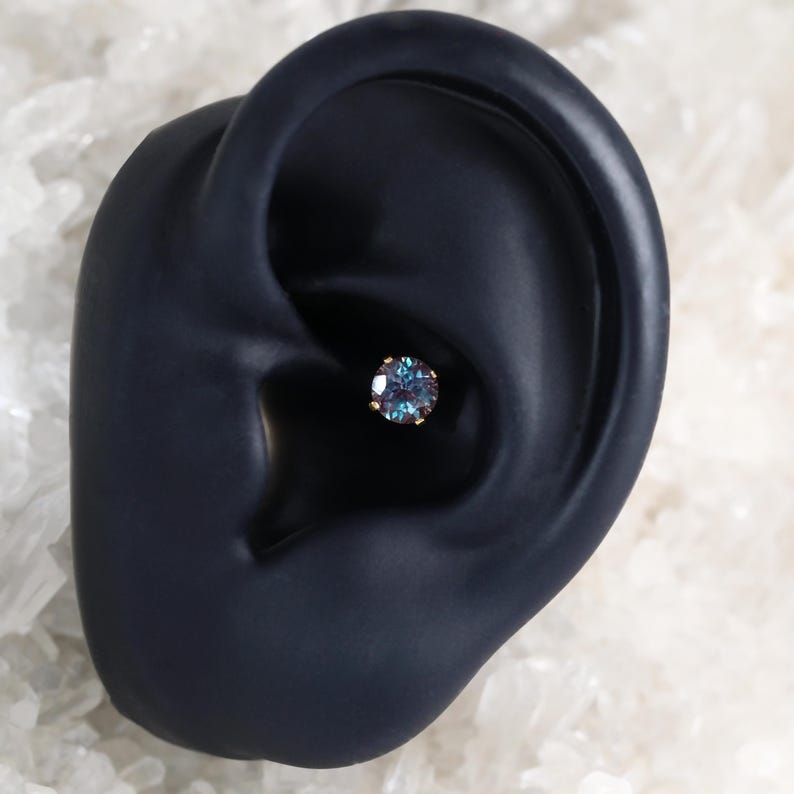 May include: A close-up of a blue gemstone earring with a gold setting. The earring is worn in a black ear model.