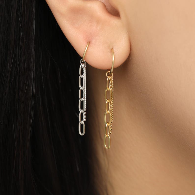 May include: A pair of dangling earrings. One earring is silver-toned with a chain design and small embellishments. The other earring is gold-toned with a chain design. Both earrings are attached to small gold hoops.