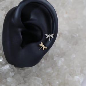 Pu&ograve; includere: Due orecchini a forma di libellula con accenti di cristallo trasparente. Gli orecchini sono mostrati su un modello di orecchio nero.