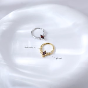 May include: Two rings, one silver and one gold, each featuring a central garnet-colored gemstone in a diamond shape. The gold ring is accented with small, clear moissanite stones. The rings are displayed on a white, reflective surface.