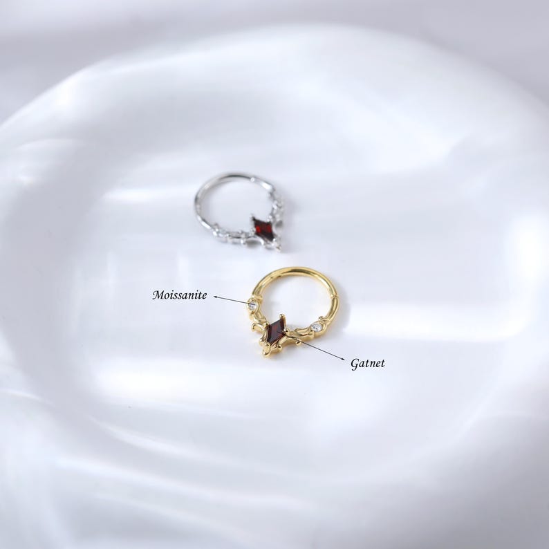 May include: Two rings, one silver and one gold, each featuring a central garnet-colored gemstone in a diamond shape. The gold ring is accented with small, clear moissanite stones. The rings are displayed on a white, reflective surface.