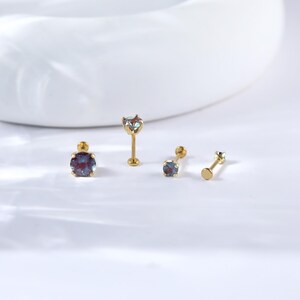 May include: Three gold body jewellery pieces with iridescent blue gemstones. The pieces are a labret stud, a barbell, and a flat back stud.