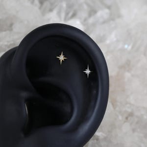 May include: Two small star-shaped earrings with a single clear stone in the centre of each star. The earrings are shown on a black ear model.