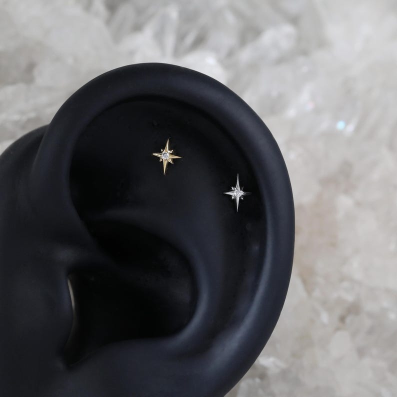 May include: Two small star-shaped earrings with a single clear stone in the centre of each star. The earrings are shown on a black ear model.