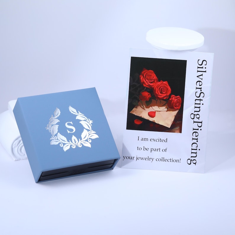 May include: A blue jewellery box with a silver 'S' in a wreath design. The box is sitting on a white surface. A white card with the text "SilverSting Piercing" and a red rose illustration is in the background. The card reads "I am excited to be part of your jewellery collection!"