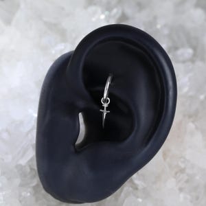 May include: A silver hoop earring with a small dagger charm. The earring is shown on a dark ear model against a white, textured background. The dagger charm has a pointed blade and a decorative handle.