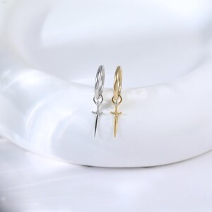 May include: Two hoop earrings, one silver-toned and one gold-toned, each with a small sword pendant. The earrings are displayed on a white surface, showcasing their intricate design and metallic finish. The swords have a small gem.