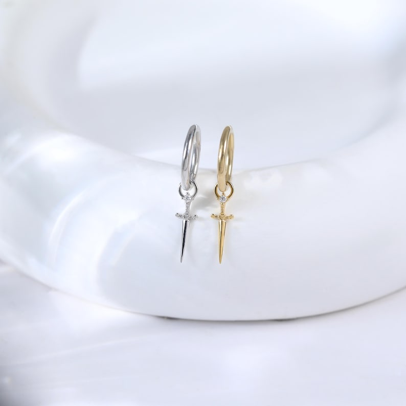 May include: Two hoop earrings, one silver-toned and one gold-toned, each with a small sword pendant. The earrings are displayed on a white surface, showcasing their intricate design and metallic finish. The swords have a small gem.