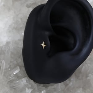 May include: A gold star-shaped earring with a small clear diamond in the centre. The earring is shown on a black ear model.
