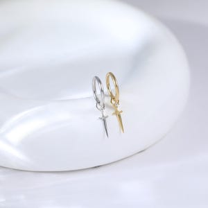 May include: A pair of hoop earrings, one silver-toned and one gold-toned, each featuring a small sword pendant. The earrings are displayed on a white, curved surface, showcasing their design and metallic finish. The sword pendants add a unique touch to the classic hoop style.