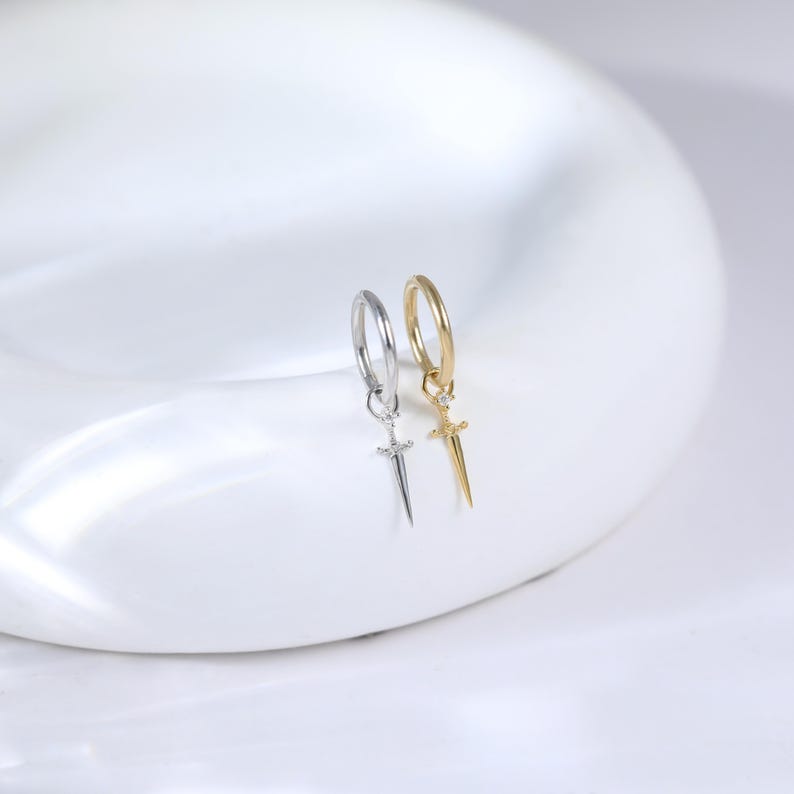 May include: A pair of hoop earrings, one silver-toned and one gold-toned, each featuring a small sword pendant. The earrings are displayed on a white, curved surface, showcasing their design and metallic finish. The sword pendants add a unique touch to the classic hoop style.