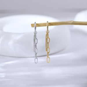 May include: Two dangle earrings, one silver and one gold, suspended from a wooden dowel. The silver earring features a chain design with a row of small, sparkling stones. The gold earring has a similar chain design.